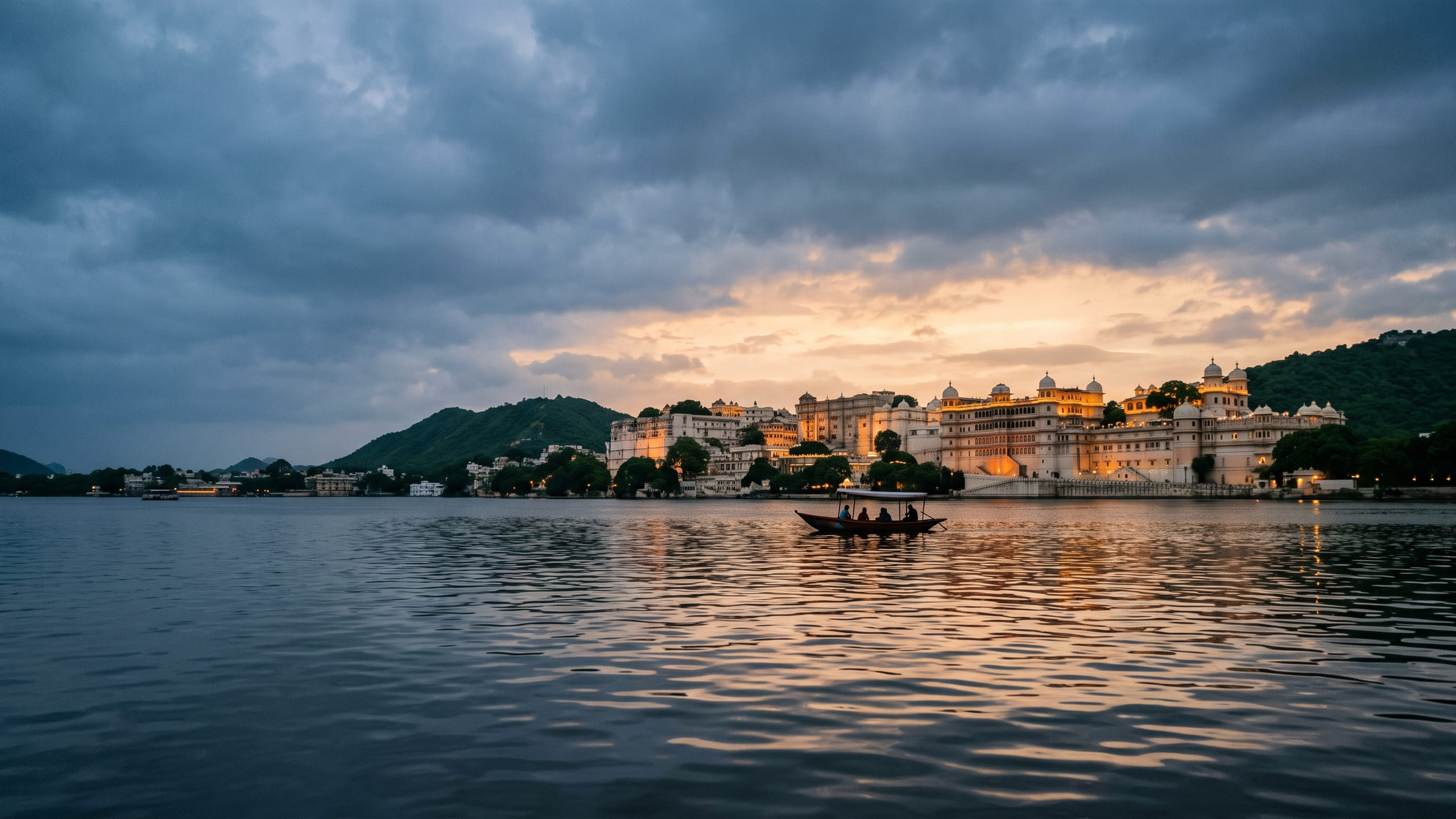 Sunset over Lake Pichola with a small boat on the water and the City Palace lit gold along the far shore under monsoon cloud